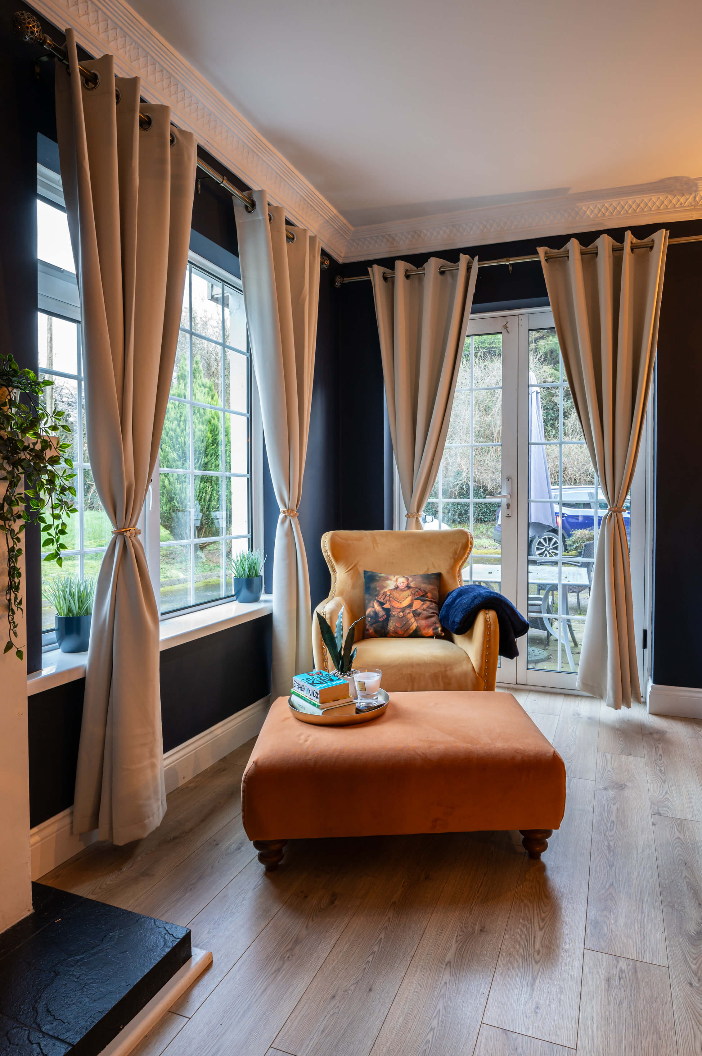 corner of a bright living room with french doors and white framed window. The walls are deep navy. The curtains are a neutral colour with a brushed gold curtain pole. A deep yellow accent chair and orange footstool are in the corner